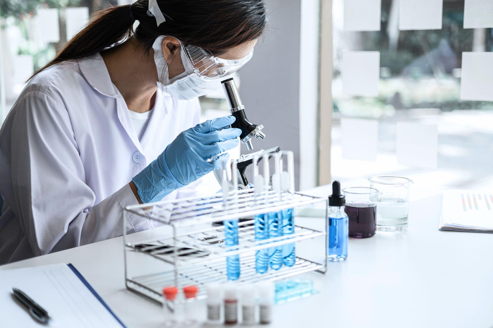 Laboratory scientist in PPE examining samples under a microscope next to racks of blue assay tubes, illustrating hit-to-lead screening activities.