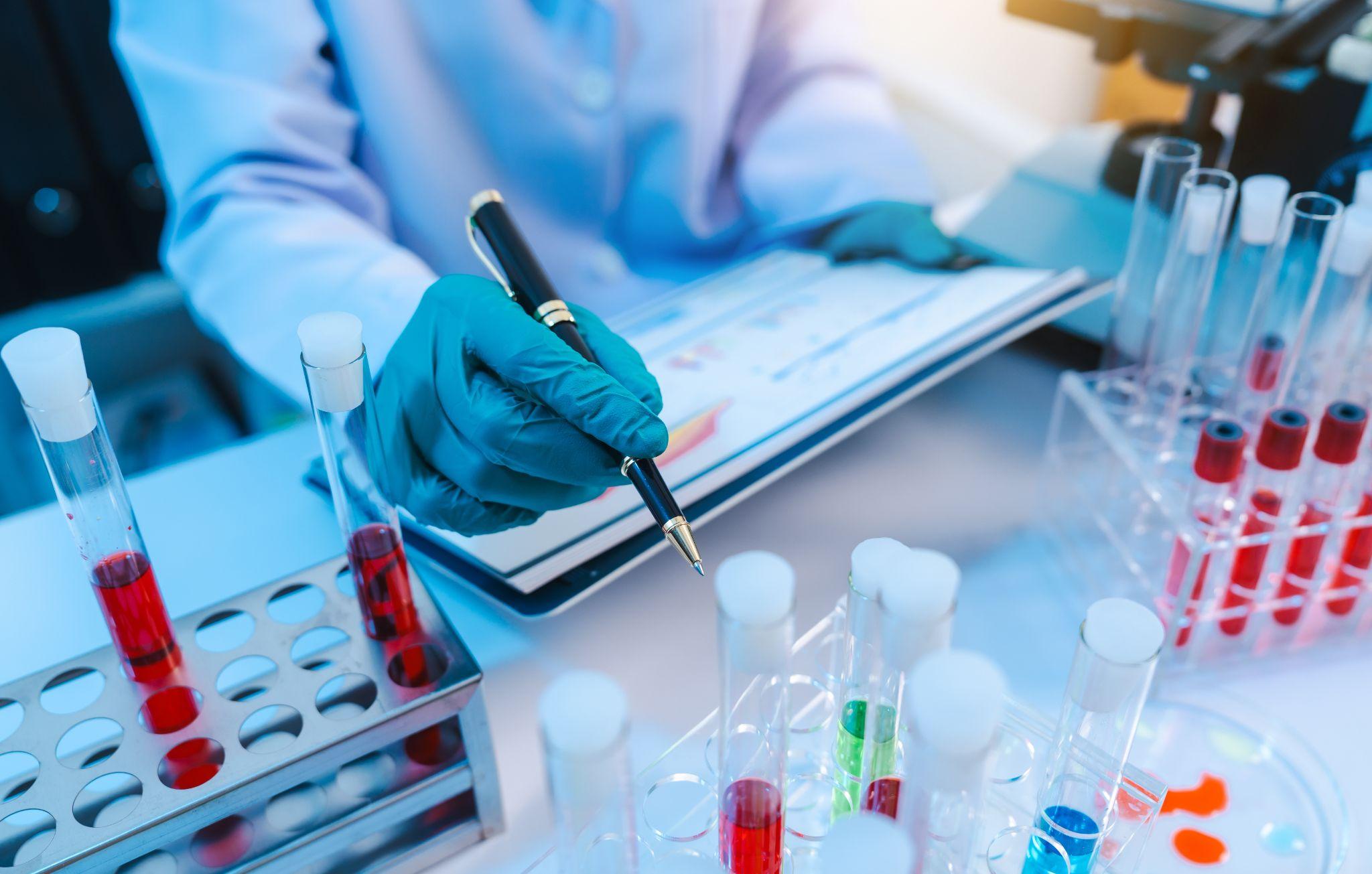 Gloved scientist pointing with a pen at assay results beside test tubes and a tablet, representing data-driven hit-to-lead decision making.