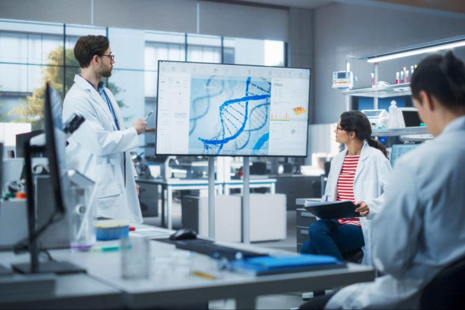 Researchers in a lab reviewing a DNA helix and data on a large monitor during a genetic-engineering planning session.