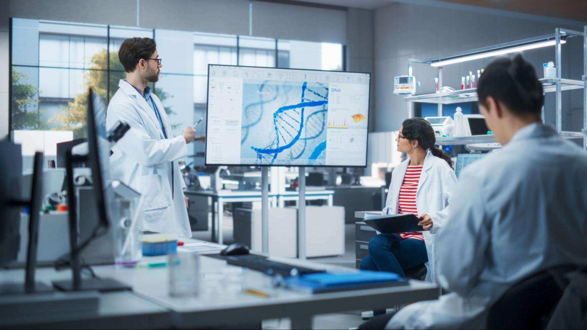 Researchers in a lab reviewing a DNA helix and data on a large monitor during a genetic-engineering planning session.