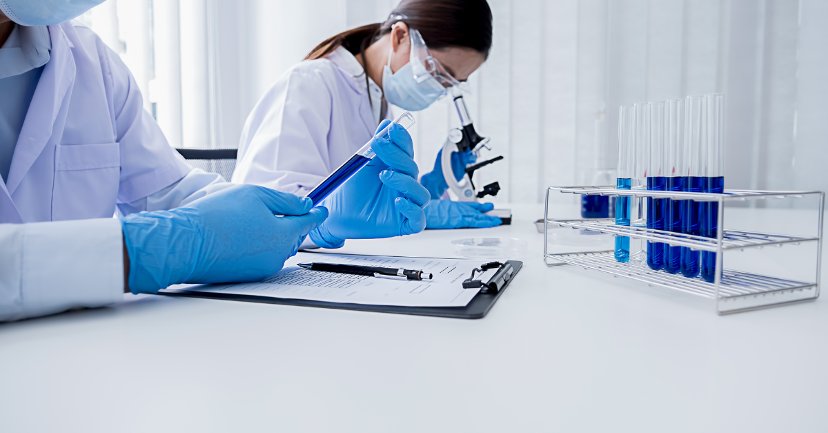 A scientist looking into a test tube while their colleague writes notes on a clipboard